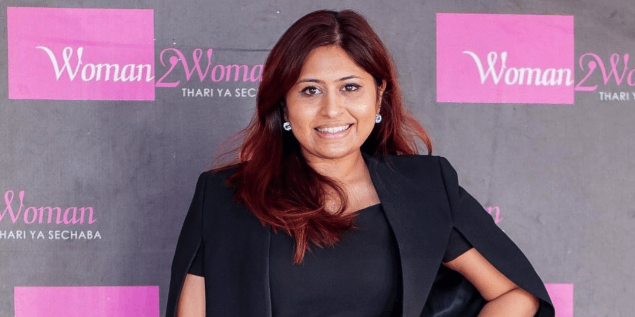 Taazima Kala-Essack, an Asian woman with chestnut coloured hair, smiles at the camera. She wears a black jacket and top. Behind her is a grey background with obscured logos in pink